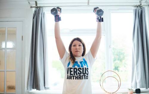 Jade holding two weights defiantly above her head wearing a Versus Arthritis t-shirt