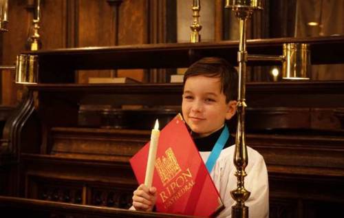 Smiling William standing in Rippon cathedral pews holding a candle and wearing chorister's robe