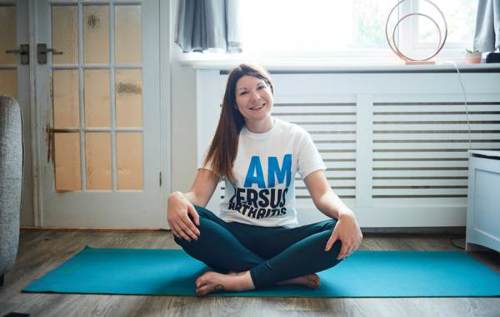 Smiling Jade wearing Versus Arthritis t-shirt and gym leggings sat on a yoga mat